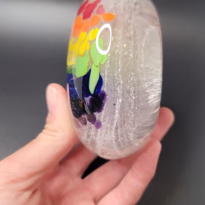 Hand holding an oval glass Rainbow Bridge paperweight featuring vibrant red, orange, yellow, green, blue, and purple color segments on one side, with swirling cremation ash trails suspended throughout the clear glass.