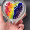 Hand holding a clear glass heart-shaped cremation paperweight featuring a vibrant Rainbow Bridge design, with swirling red, orange, yellow, green, blue, and purple color segments suspended inside and delicate bubble patterns throughout.