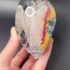 Side view of a clear glass heart-shaped Rainbow Bridge cremation paperweight, showing suspended colorful bands of blue, purple, green, and yellow on one side and sparkling white bubble trails encased within the glass, held in a person's hand.
