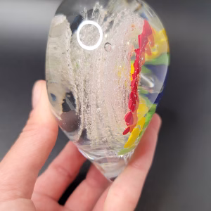 Side view of a clear glass heart-shaped Rainbow Bridge cremation paperweight, showing suspended colorful bands of blue, purple, green, and yellow on one side and sparkling white bubble trails encased within the glass, held in a person's hand.