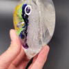 Side view of a clear glass heart-shaped Rainbow Bridge cremation paperweight, showing suspended colorful bands of blue, purple, green, and yellow on one side and sparkling white bubble trails encased within the glass, held in a person's hand.