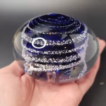 Hand holding a clear glass memorial paperweight featuring deep blue swirling bands encased inside, with cremation ash forming fine white trails and speckles layered throughout the design.