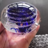 Hand holding a clear glass memorial paperweight featuring deep blue swirling bands encased inside, with cremation ash forming fine white trails and speckles layered throughout the design.