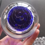 Top view of a clear glass memorial paperweight held in a hand, featuring a deep blue core with swirling rings encased inside and cremation ash forming delicate white speckled trails around the center.