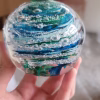 Hand holding a clear glass memorial paperweight with layered blue and green swirling bands encased inside, featuring cremation ash suspended in delicate white trails and bubbles throughout the design.
