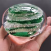 Hand holding a clear glass memorial paperweight viewed from the side, showcasing vibrant green swirling bands encased inside with cremation ash forming delicate white trails layered between the colored glass ribbons.
