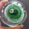 Top view of a clear glass memorial paperweight held in a hand, featuring vibrant green swirling bands encased inside, with cremation ash forming delicate white speckled patterns concentrated near the center and radiating outward.