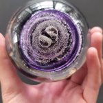 Hand holding a clear glass memorial paperweight viewed from the top, showing deep purple swirling bands encased inside with a dense pattern of suspended cremation ash concentrated toward the center.