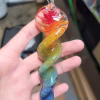 A 6-inch rainbow glass spinner infused with cremation ash, held in a hand. The piece features a clear glass loop at the top and a flowing twisted design with vibrant red, orange, yellow, green, and blue swirls running down its length.