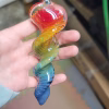 A 6-inch rainbow glass spinner infused with cremation ash, held in a hand. The piece features a clear glass loop at the top and a flowing twisted design with vibrant red, orange, yellow, green, and blue swirls running down its length.