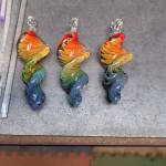 Three 3-inch rainbow glass spinners infused with cremation ash, arranged side by side on a workbench. Each spinner features swirling red, orange, yellow, green, and blue colors with a twisted sculptural shape and a clear glass loop at the top.