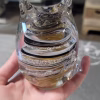 Hand holding a clear glass cat-shaped paperweight with sculpted ears and a curled tail, featuring layered black, tan, and cream swirling bands inside. Cremation ash forms delicate white speckles suspended throughout the spiral design.