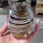 Hand holding a clear glass cat-shaped paperweight with sculpted ears and a curled tail, featuring layered black, tan, and cream swirling bands inside. Cremation ash forms delicate white speckles suspended throughout the spiral design.