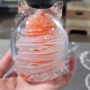 Hand holding a clear glass cat-shaped paperweight featuring swirling orange and white bands encased inside, with cremation ash suspended in fine sparkling trails throughout the design. The sculpted ears and curled tail create a stylized cat silhouette.