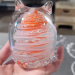 Hand holding a clear glass cat-shaped paperweight featuring swirling orange and white bands encased inside, with cremation ash suspended in fine sparkling trails throughout the design. The sculpted ears and curled tail create a stylized cat silhouette.