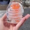 Hand holding a clear glass cat-shaped paperweight featuring swirling orange and white bands encased inside, with cremation ash suspended in fine sparkling trails throughout the design. The sculpted ears and curled tail create a stylized cat silhouette.