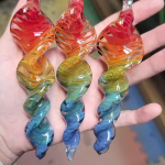 Three 6-inch rainbow glass spinner ornaments infused with cremation ash, featuring vibrant red, orange, yellow, green, and blue swirled patterns, displayed resting on a hand