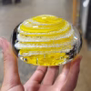 Hand holding a clear glass memorial paperweight featuring vibrant yellow swirling bands encased inside, with cremation ash suspended in delicate white trails throughout the design.