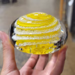 Hand holding a clear glass memorial paperweight featuring vibrant yellow swirling bands encased inside, with cremation ash suspended in delicate white trails throughout the design.