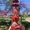 A hand holding a red glass lighthouse infused with cremation ash, featuring a spiraling red core with shimmering white ash suspended throughout. The lighthouse is photographed outdoors against a bright blue sky, with blooming pink trees and soft greenery blurred in the background.