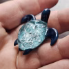 Handcrafted glass sea turtle cremation pendant with deep blue flippers and a textured aqua shell containing white flecks of encased cremation ashes, shown resting in the hand to display size and detail