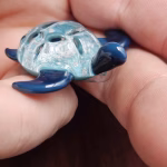 Handcrafted glass sea turtle cremation pendant shown from a low side angle, featuring deep blue flippers and a domed aqua shell infused with white flecks of encased cremation ashes, held between fingers to show size and sculpted detail