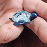 Handcrafted glass sea turtle cremation pendant shown from a low side angle, featuring deep blue flippers and a textured aqua shell with white flecks of encased cremation ashes, held between fingers for scale and detail