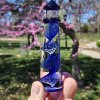 A hand holding a cobalt-blue glass lighthouse infused with cremation ash, featuring a spiral core of swirling blue and white bubbles. The lighthouse is photographed outdoors with blooming pink trees and bright spring greenery softly blurred in the background.