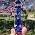 A hand holding a cobalt-blue glass lighthouse infused with cremation ash, featuring a spiral core of swirling blue and white bubbles. The lighthouse is photographed outdoors with blooming pink trees and bright spring greenery softly blurred in the background.