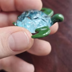Handcrafted glass sea turtle cremation pendant shown from the side, featuring smooth green flippers and a domed aqua shell with white flecks of encased cremation ashes, held between fingers to show detail and scale