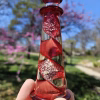 A hand holding a red glass lighthouse infused with cremation ash, featuring a spiraling red core with shimmering white ash suspended throughout. The lighthouse is photographed outdoors against a bright blue sky, with blooming pink trees and soft greenery blurred in the background.