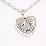 Close-up image of a heart-shaped stainless steel cremation pendant featuring two raised baby feet on the front, shown on a silver chain against a white background.