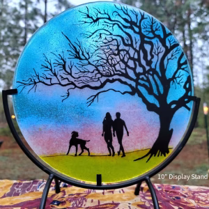 Round glass memorial sun plate featuring a silhouetted couple walking with their dog beneath a large branching tree, set against a blue, pink, and yellow gradient background and displayed on a 10-inch metal stand.