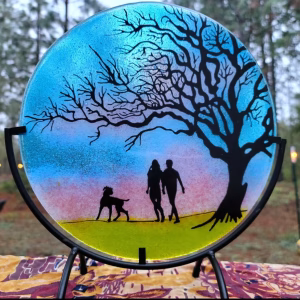 Round glass memorial sun plate featuring a silhouetted couple walking with their dog beneath a large branching tree, set against a blue, pink, and yellow gradient background and displayed on a 10-inch metal stand.