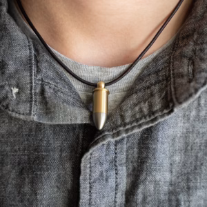 Model wearing a bullet-shaped cremation pendant with a brass-toned body and stainless steel tip, hung on a dark cord necklace and shown against a casual gray shirt.