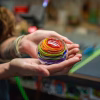 Hands gently holding a round glass paperweight with vibrant rainbow-colored swirling bands and suspended cremation ash inside, shown in a warm studio setting.
