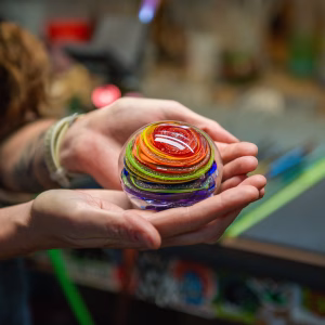 Hands gently holding a round glass paperweight with vibrant rainbow-colored swirling bands and suspended cremation ash inside, shown in a warm studio setting.