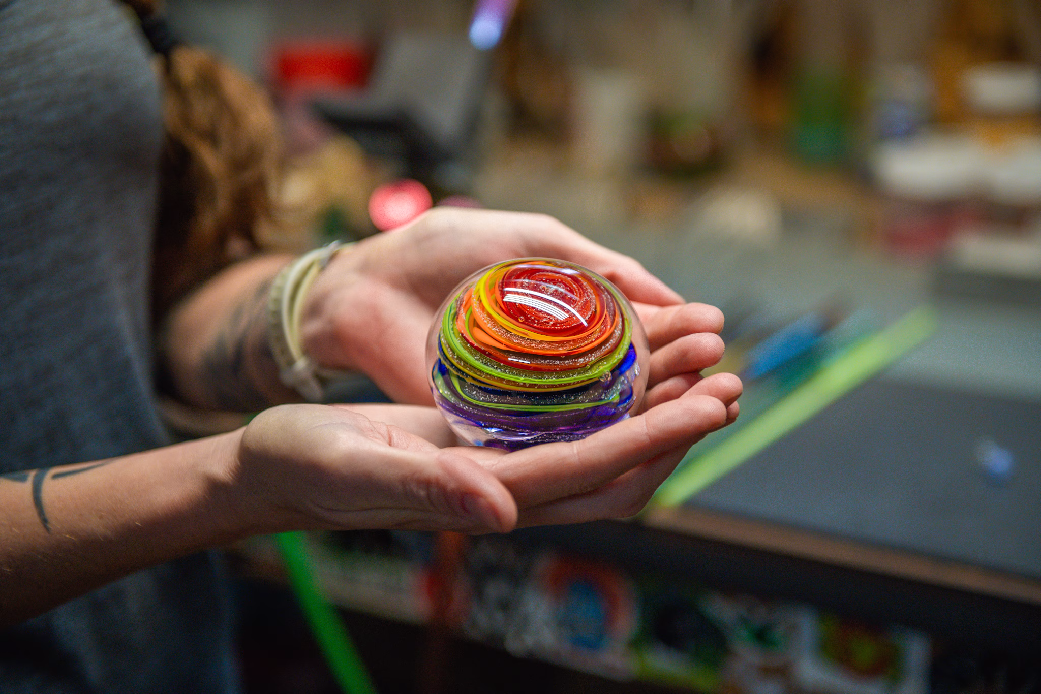 Hands gently holding a round glass paperweight with vibrant rainbow-colored swirling bands and suspended cremation ash inside, shown in a warm studio setting.