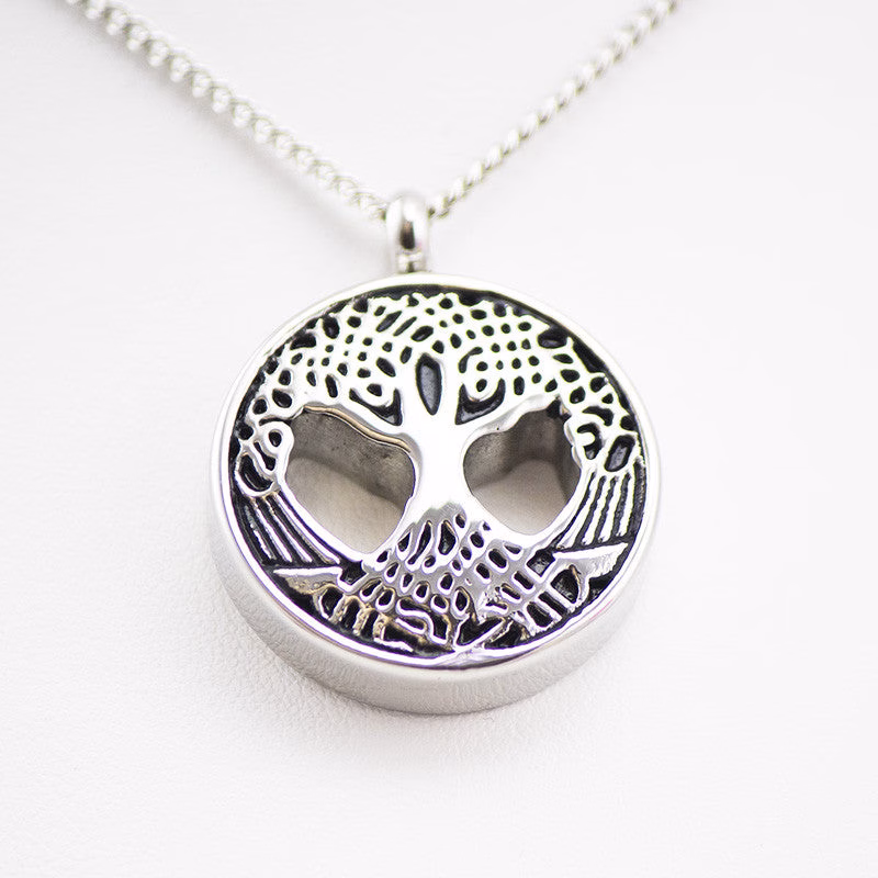 Silver round pendant with an intricate tree design on the front, featuring cutout heart-shaped openings and detailed roots, photographed against a white background.