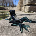 A dark green glass frog sculpture infused with cremation ash, posed on a sunlit stone ledge. The frog is viewed from a low side angle, highlighting its outstretched translucent green legs and the shimmering ash embedded along its back. A pond and stone wall fade softly into the background.