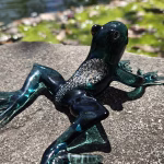 A dark green glass frog sculpture infused with cremation ash, shown from a rear angled view as it rests on a textured stone surface near a pond. Sunlight reveals the shimmering ash pattern along its back and the translucent green legs stretched outward.