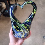 Hand holding a glass heart-shaped cremation sculpture featuring swirling blue, yellow, and black colors throughout, with cremation ash suspended in the base and fluid glass curves forming an open heart shape.