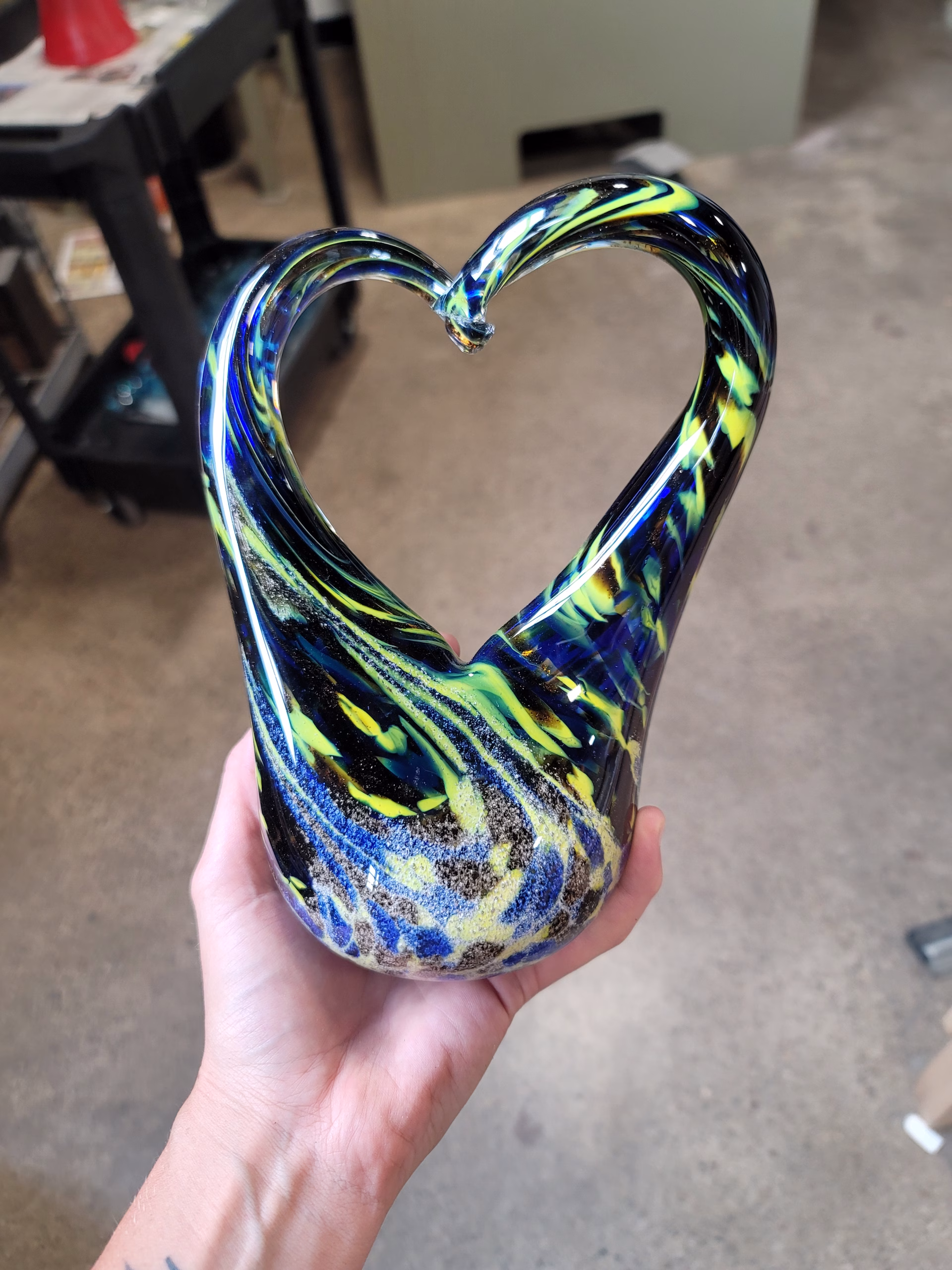 Hand holding a glass heart-shaped cremation sculpture featuring swirling blue, yellow, and black colors throughout, with cremation ash suspended in the base and fluid glass curves forming an open heart shape.
