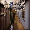 Dragonfly suncatcher with green glass body, blue head, hand-painted transparent wings, and dangling crystal prisms, hanging outdoors between buildings along a narrow walkway