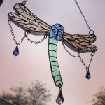 Dragonfly suncatcher with green glass body, blue head, hand-painted transparent wings, and dangling crystal prisms, hanging outdoors against the sky at sunrise