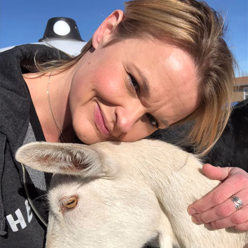 Sands of Time artist smiling while gently holding a white goat outdoors in bright sunlight on a clear day