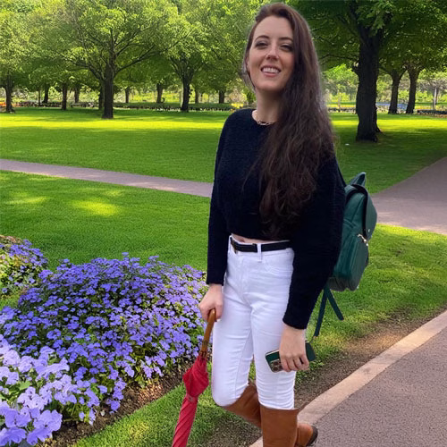 Kat, Sands of Time team member, standing in a green park walkway with purple flowers, holding a red umbrella and wearing a black sweater, white pants, and brown boots