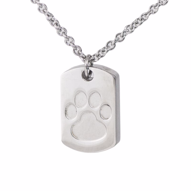 Close-up image of a stainless steel dog tag–style cremation pendant featuring an engraved paw print design on the front.
