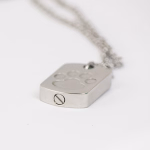 Close-up image of a stainless steel dog tag–style cremation pendant engraved with a single paw print, shown lying flat with a visible screw opening on the bottom edge and a silver chain attached.