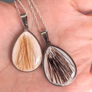 Two pear-shaped memorial pendants filled with preserved hair, displayed in an open hand, each set in a silver bezel on a delicate chain.